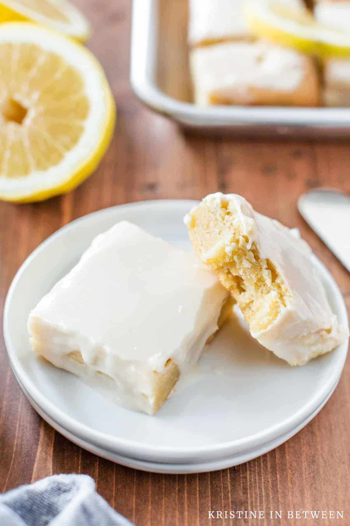 Two lemon brownies sitting on a white plate with a lemon and the rest of the bars in the background.
