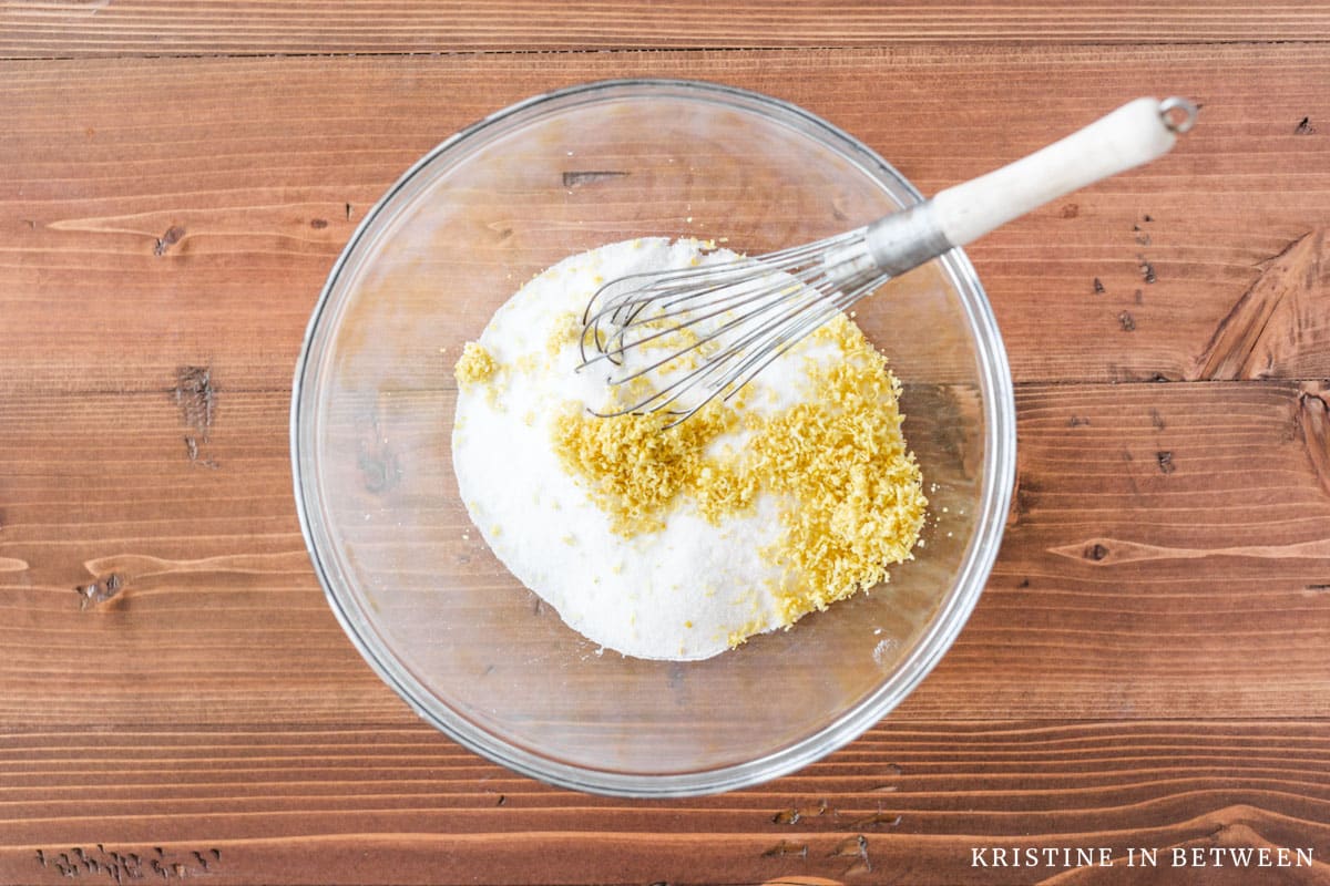 Sugar and lemon zest in a glass bowl with a whisk.