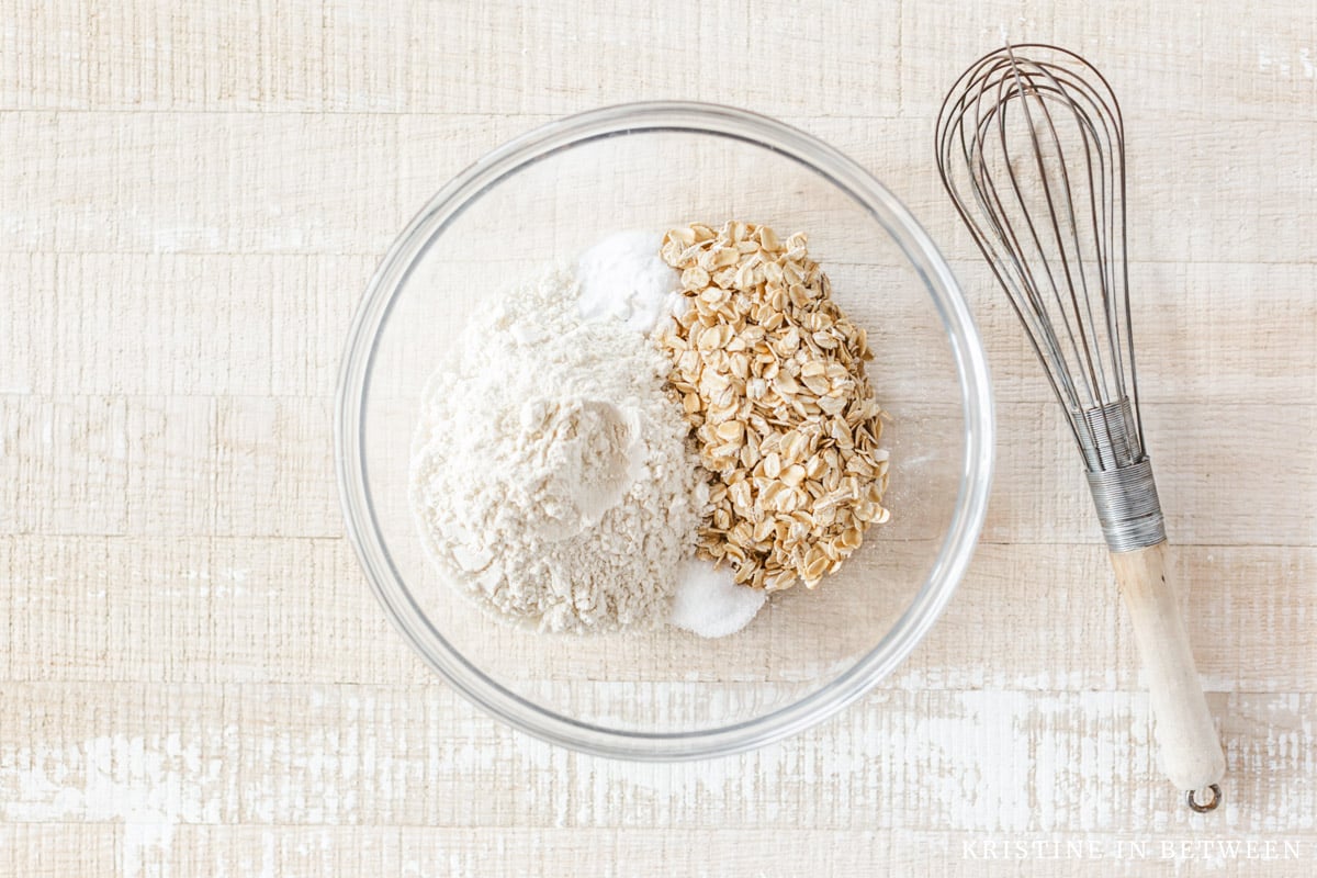 Flour, oats, salt, and baking soda in a glass bowl with a whisk.
