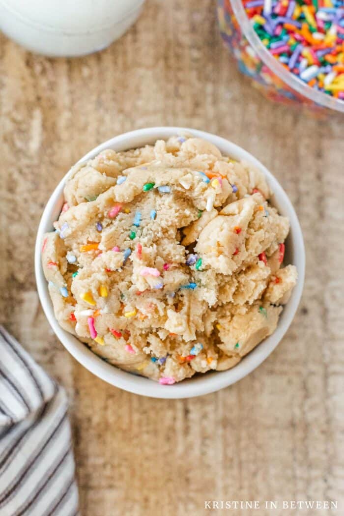 A small bowl of edible sugar cookie dough with sprinkles sitting on a wooden table.