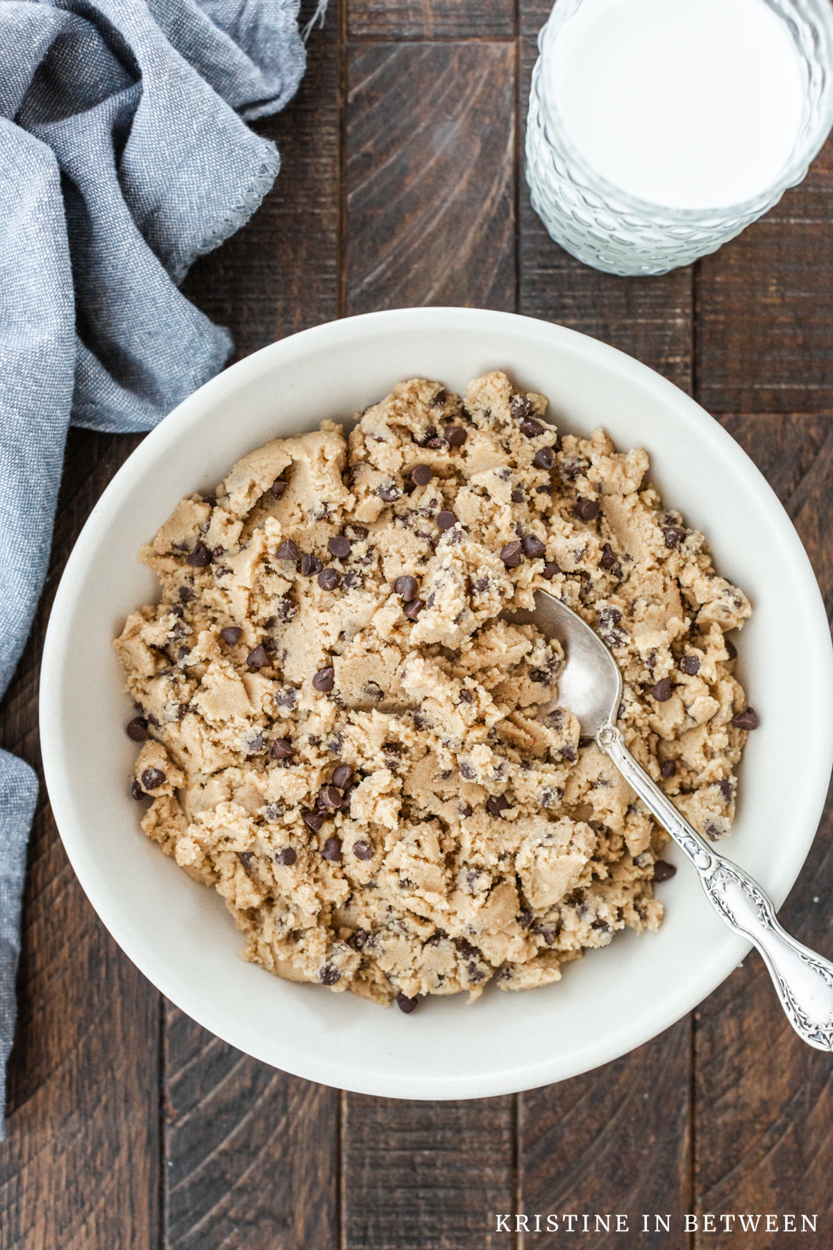 A bowl of edible cookie dough with a spoon in it and glass of milk in the background.