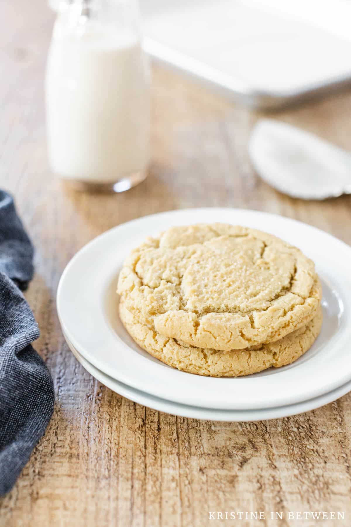 Two sugar cookie stacked up on a white plate with a spatula and a cookie sheet in the background.