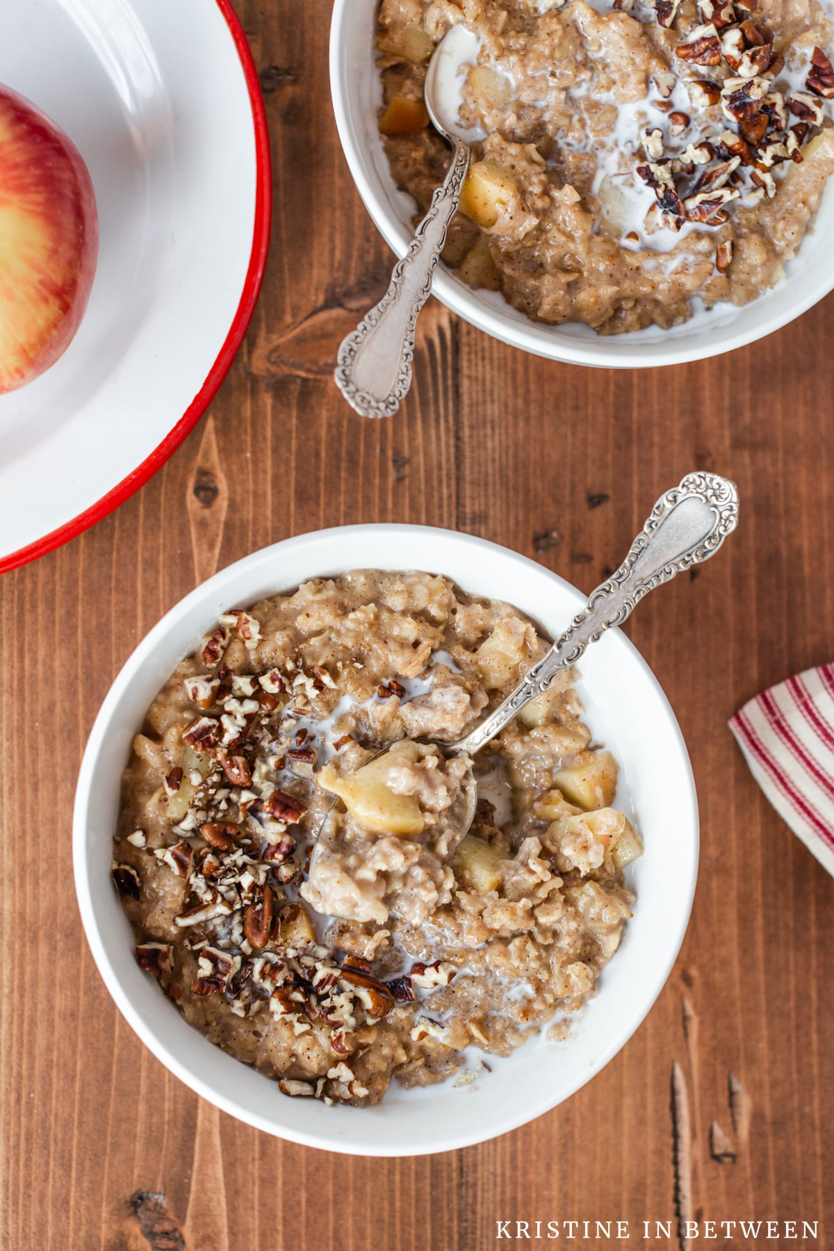 Two bowls of apple pie oatmeal with spoons in them and an apple in the background.