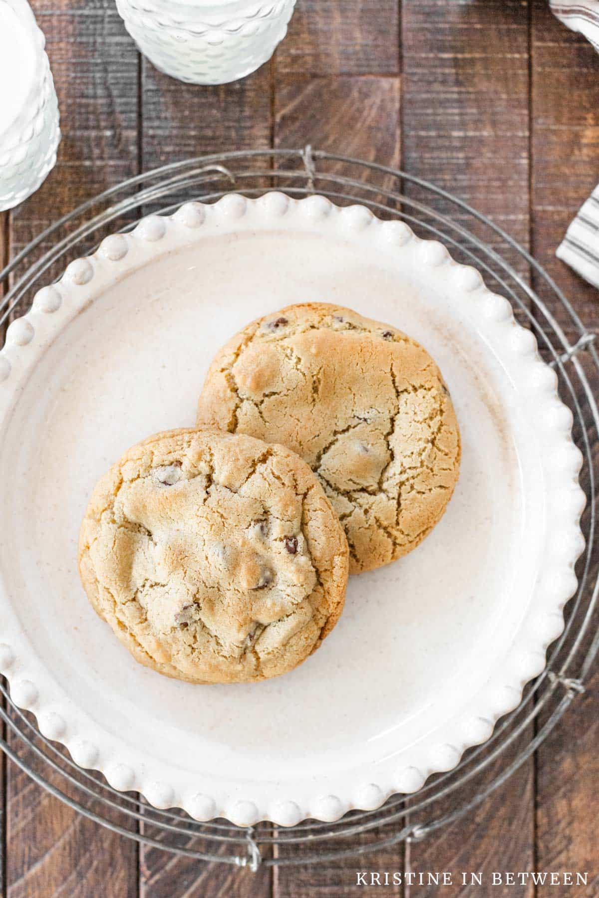 Two chocolate chip cookies sitting on a white plate with two glasses of milk in the background.