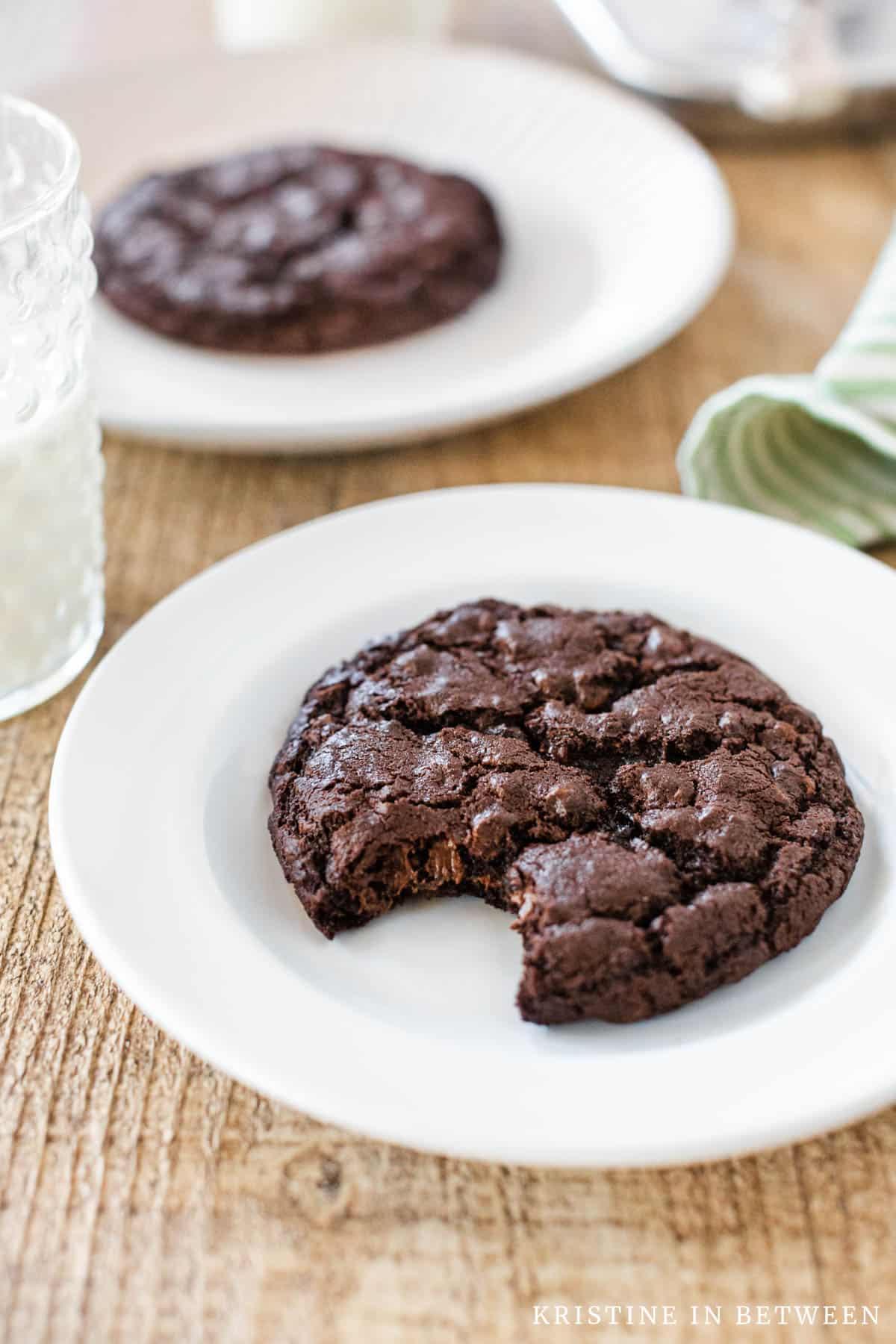 Two dark chocolate cookies sitting on white plates, one with a bite out of it.