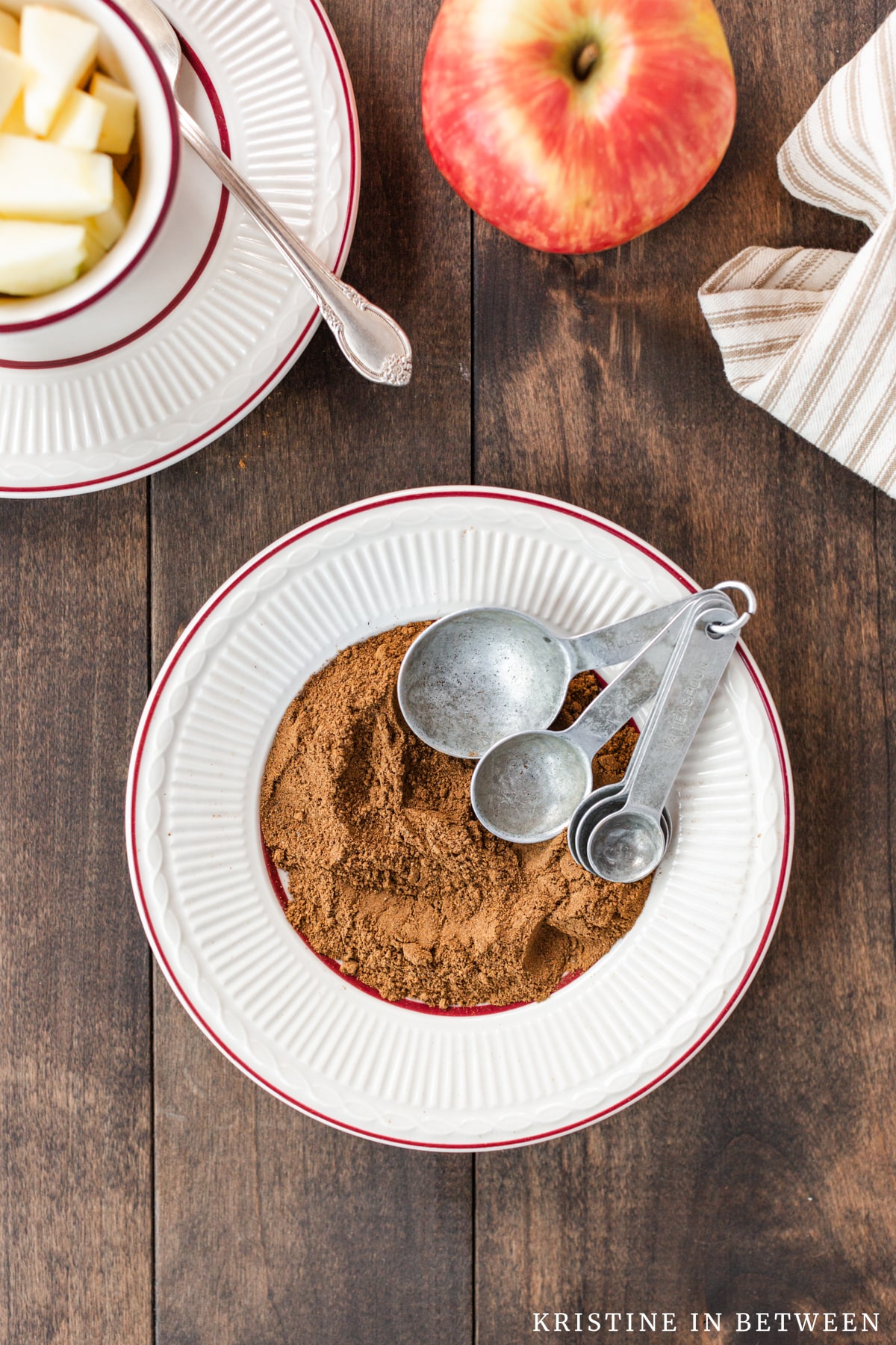 Homemade apple pie spice on a white plate with maroon trim and a small bowl of apples with a spoon in the background.