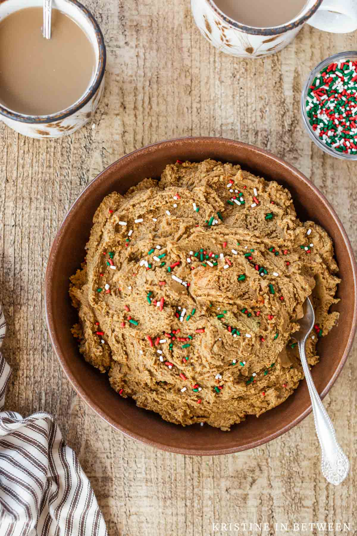 A bowl of edible gingerbread cookie dough with Christmas sprinkles in a brown bowl with a spoon.