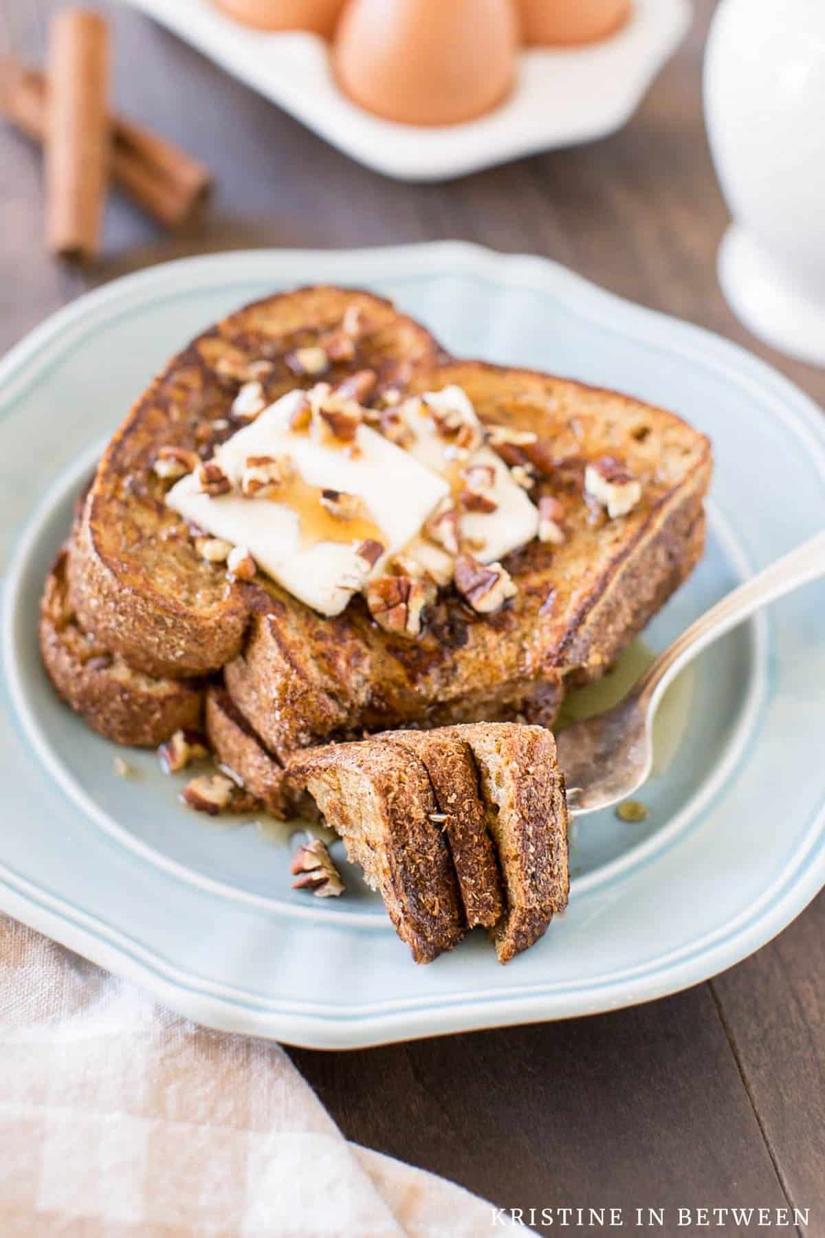 A bite of pumpkin French toast on a fork with eggs and syrup in the background.