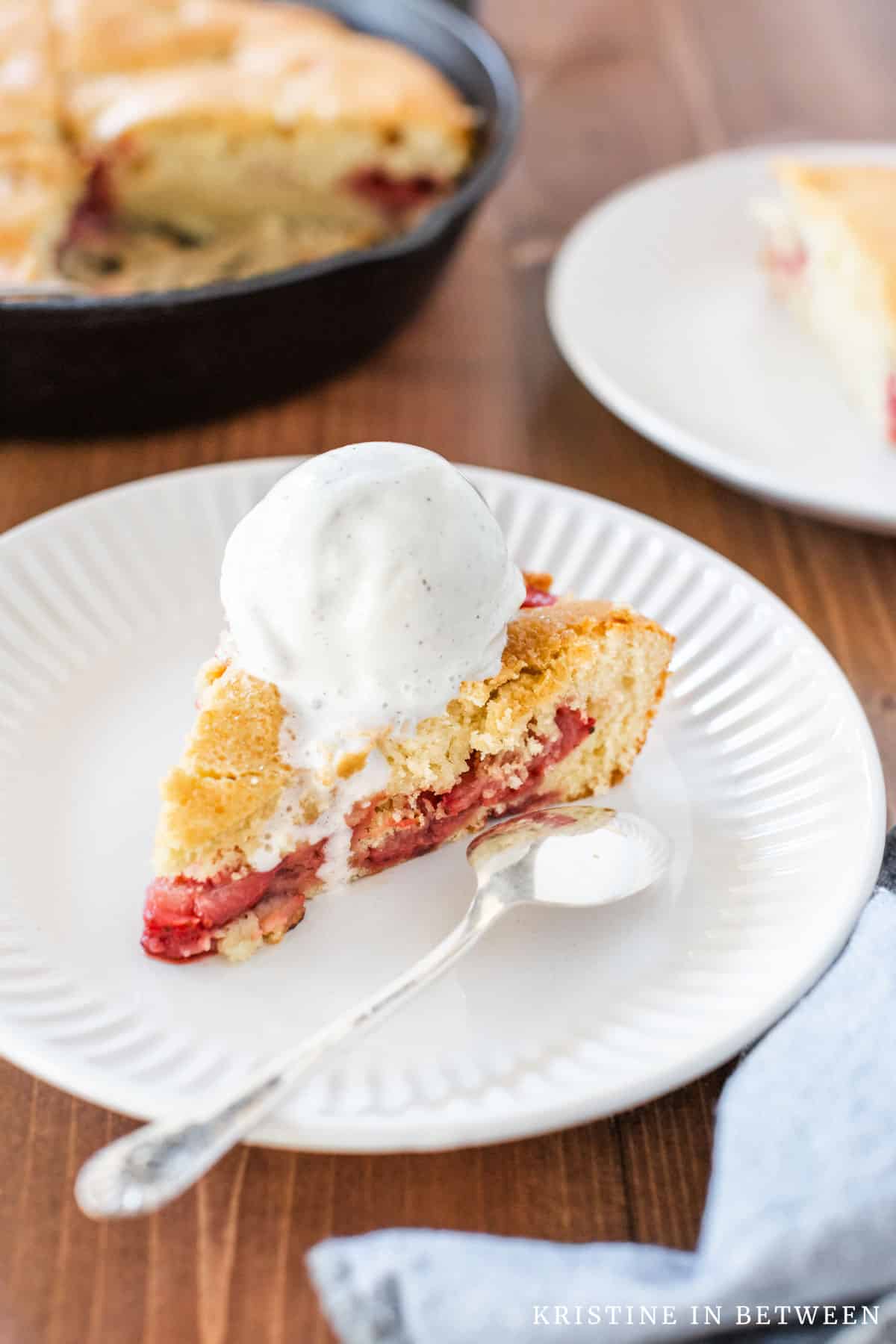 A slice of strawberry skillet cake on a white plate, topped with vanilla ice cream.