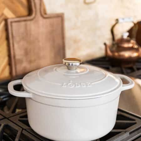 An oyster colored Lodge Dutch oven sitting on gas cook top with a copper tea kettle and two cutting boards in the background.