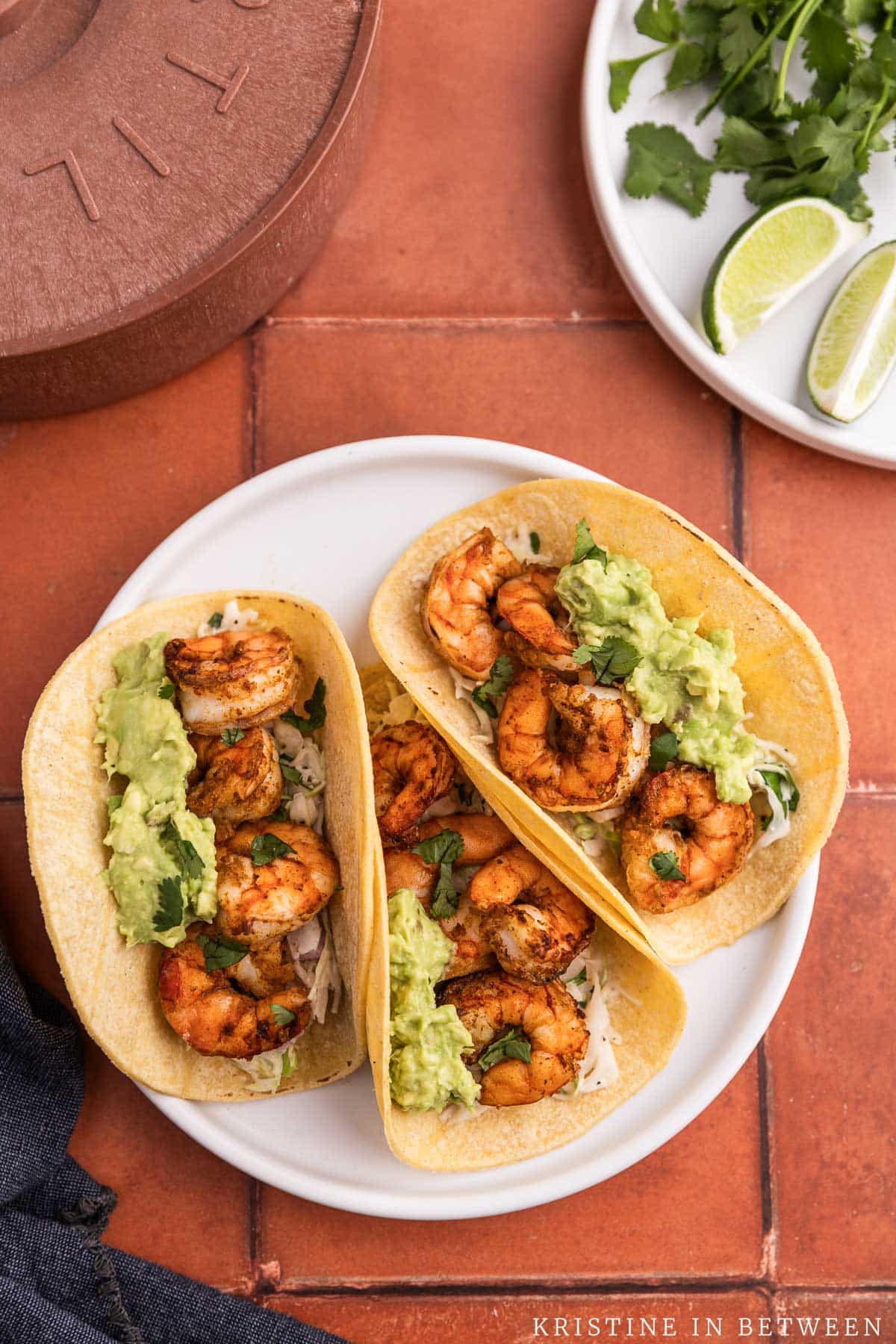 Three air fryer shrimp tacos with avocado on top sitting on a white plate.