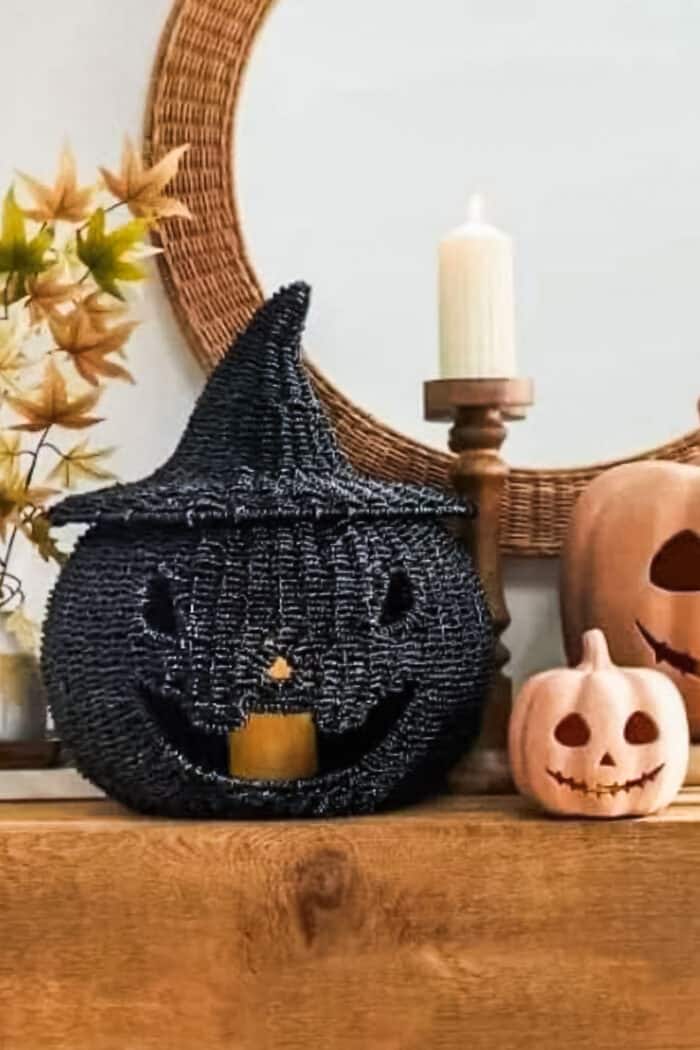A black seagrass pumpkin sitting on a mantel with two smaller pumpkins and a pillar candle.