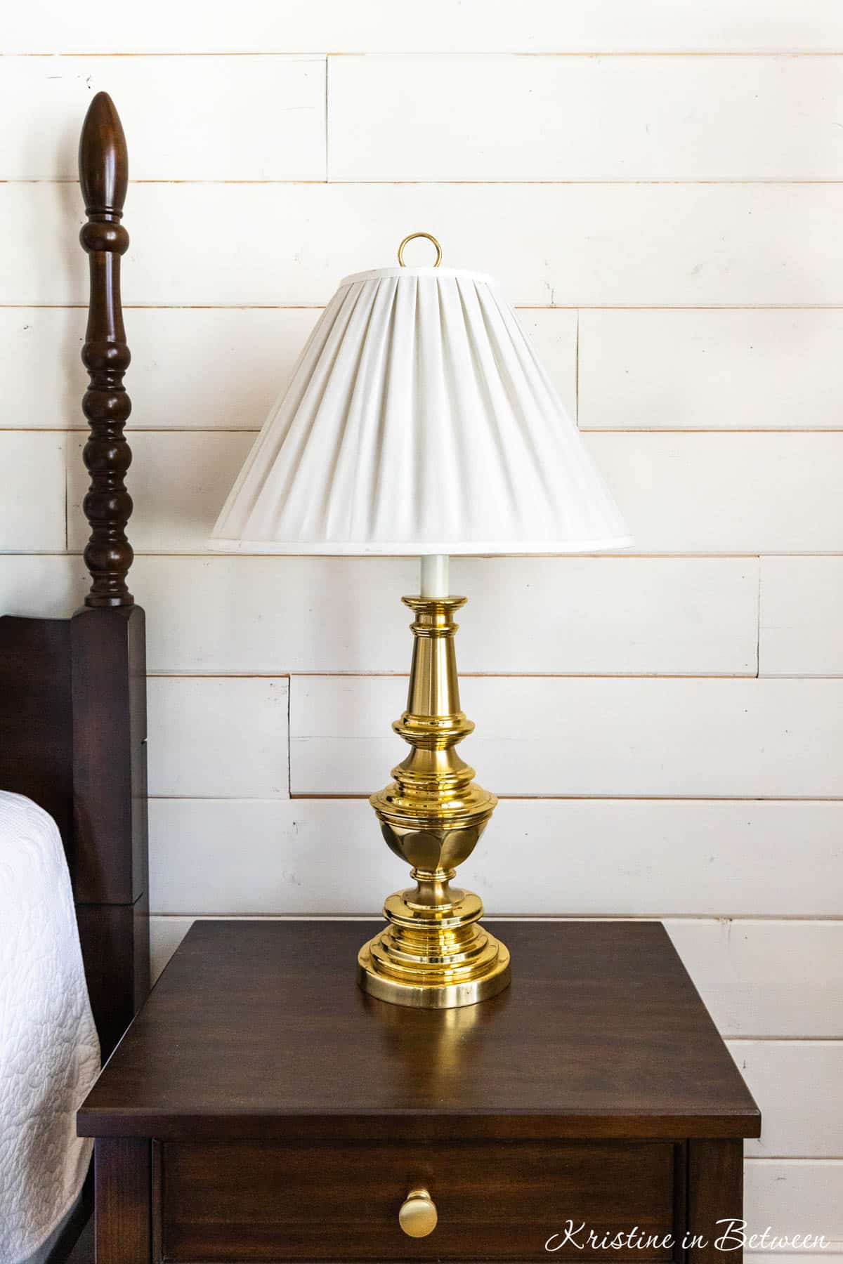 A brass Stiffel lamp sitting on a nightstand with a white pleated lamp shade.