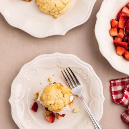 A strawberry shortcake biscuit sitting on a white scalloped plate with a fork next to it.