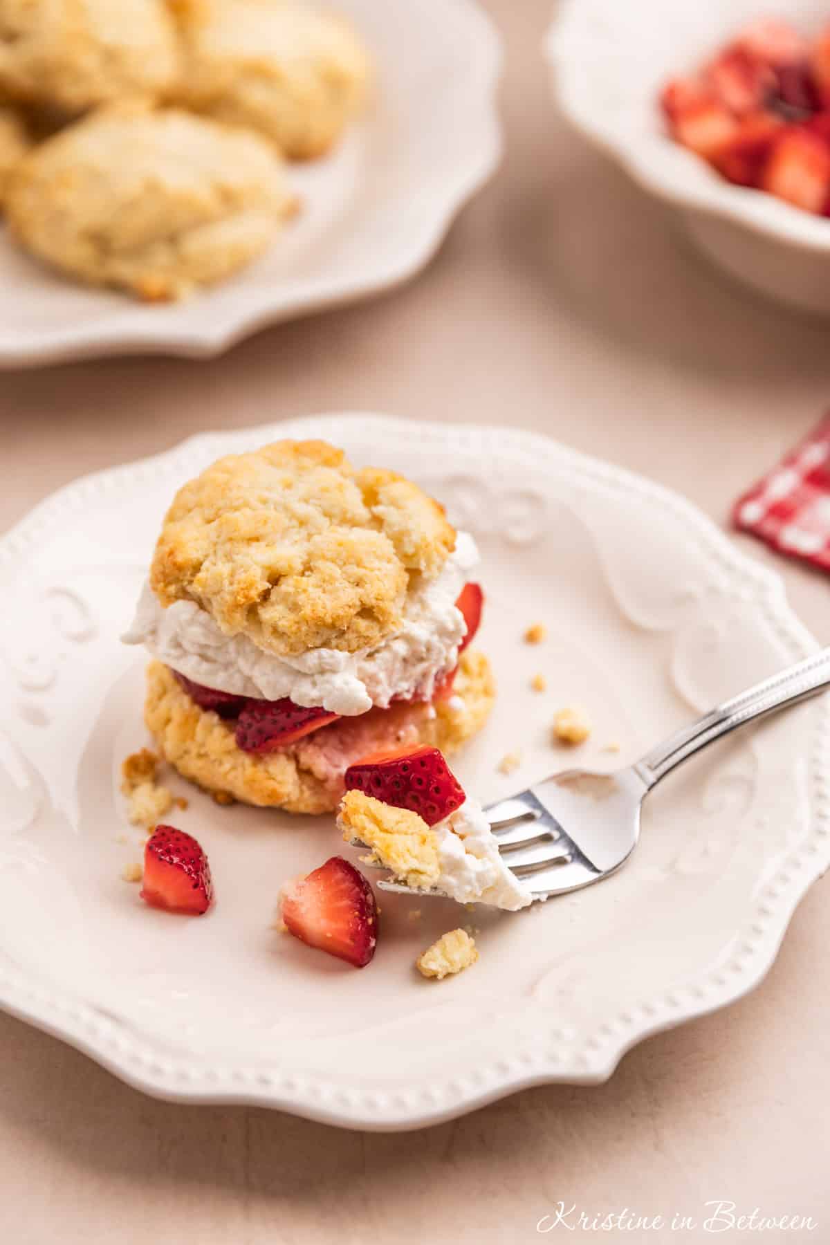 A strawberry shortcake biscuit sitting on a white plate with a bite on a fork.