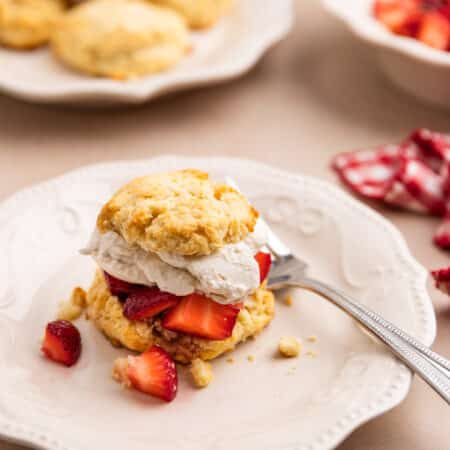 A strawberry shortcake biscuit sitting on a white scalloped plate with a fork next to it.