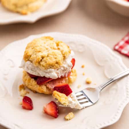 A strawberry shortcake biscuit sitting on a white plate with a bite on a fork.