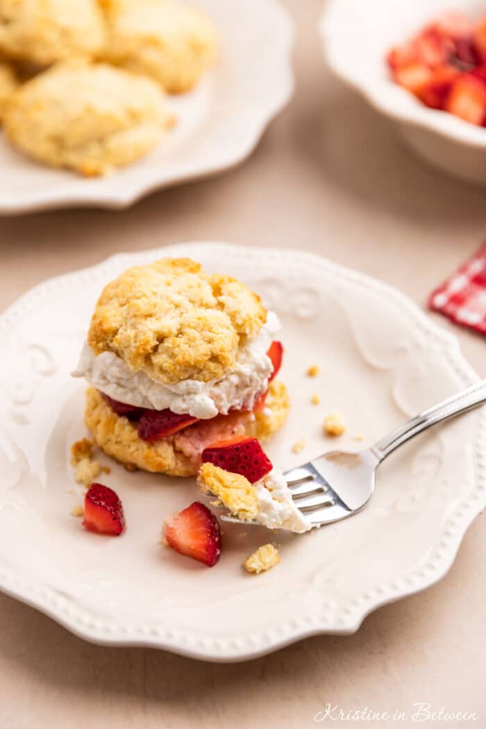 A strawberry shortcake biscuit sitting on a white plate with a bite on a fork.