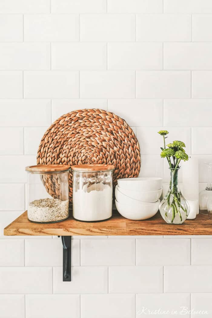 Glass jars of ingredients and a stack of bowls sitting on a wooden shelf with subway tiles in the background.