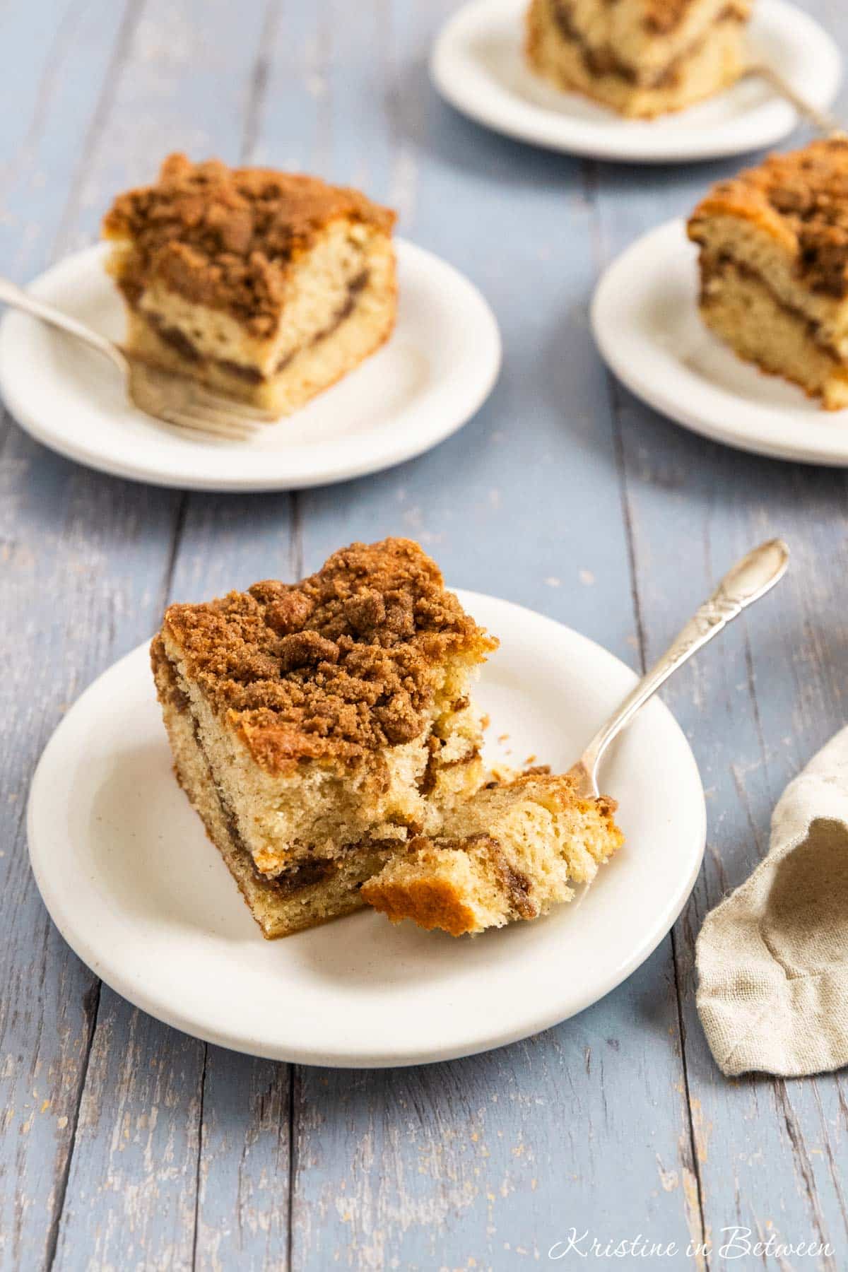 A piece of coffee cake with a bite out of it on a white plate with several other plates in the background.