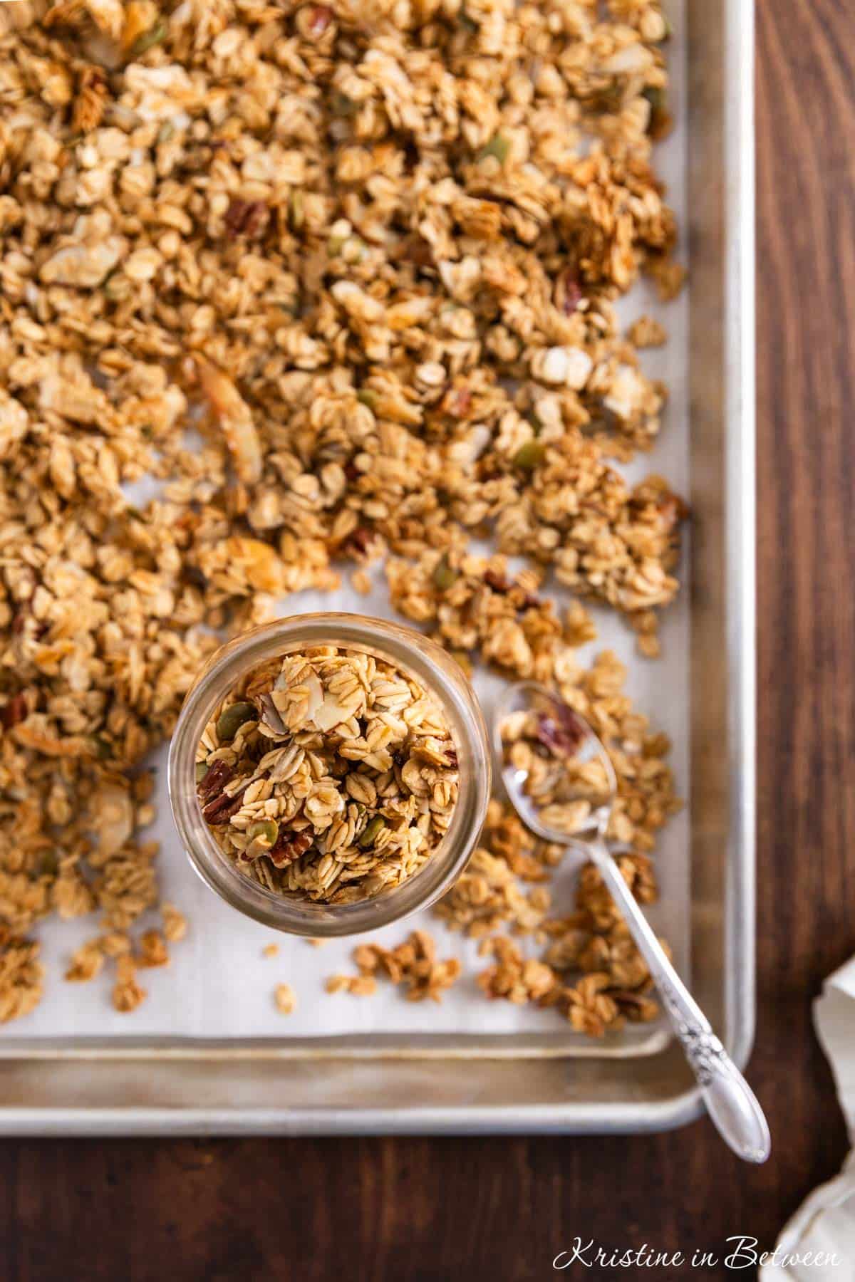 A jar of everyday granola sitting on a baking sheet of more granola with a spoon.