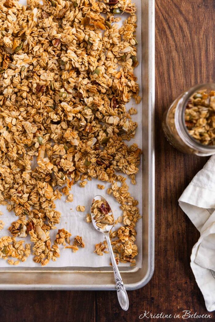 Homemade granola on an aluminum baking sheet with a spoon sitting next to it.