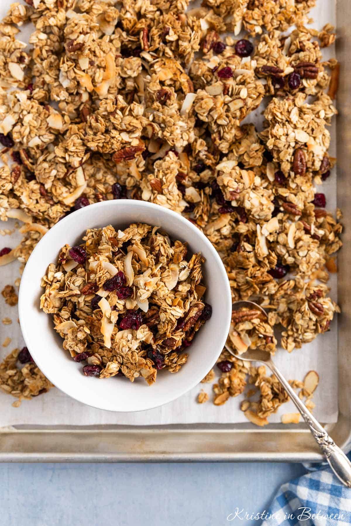 A bowl of freshly baked homemade granola sitting on a baking tray of granola with an antique spoon.
