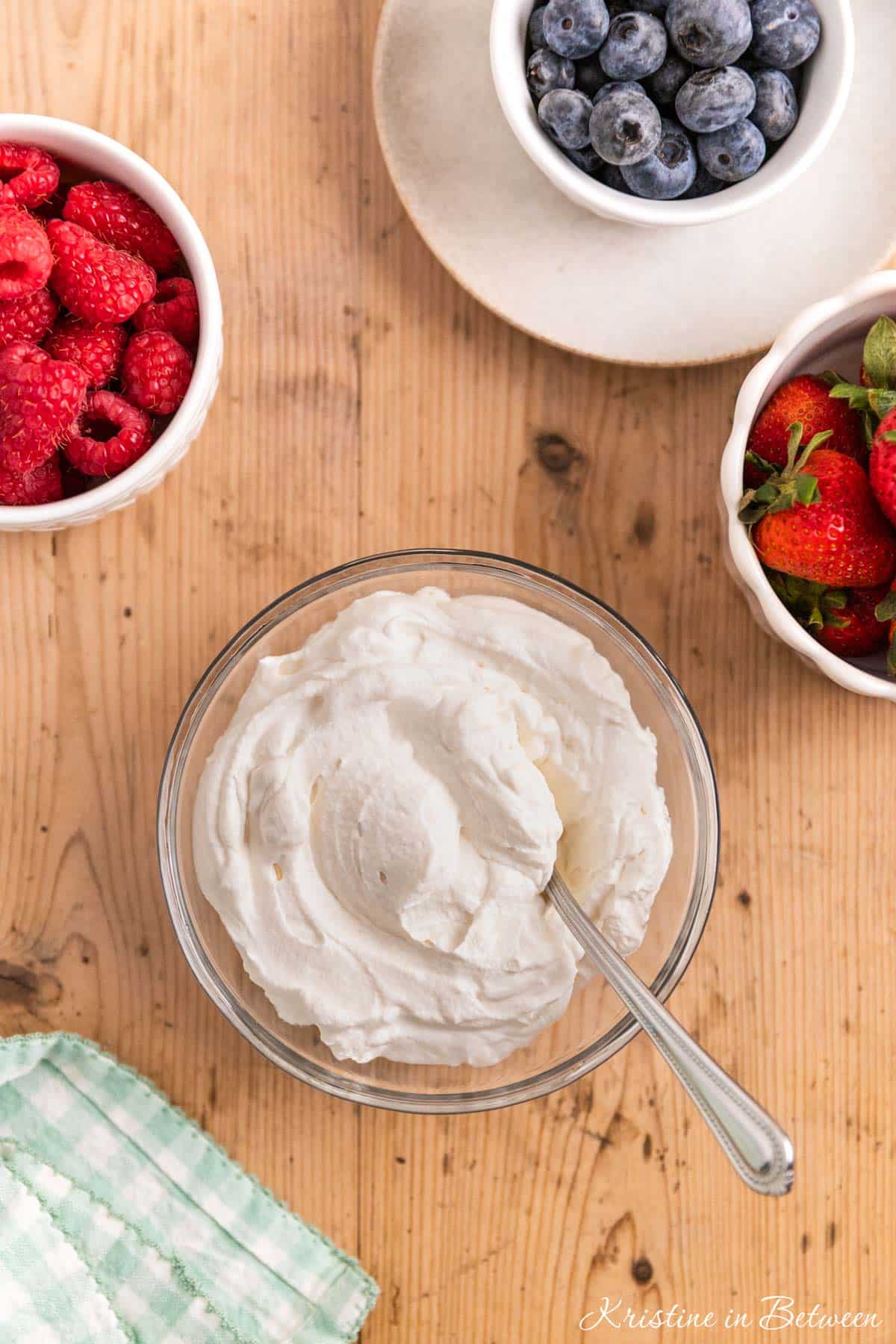 A bowl of homemade whipped cream in a glass bowl with a spoon sitting next to small bowls of berries.