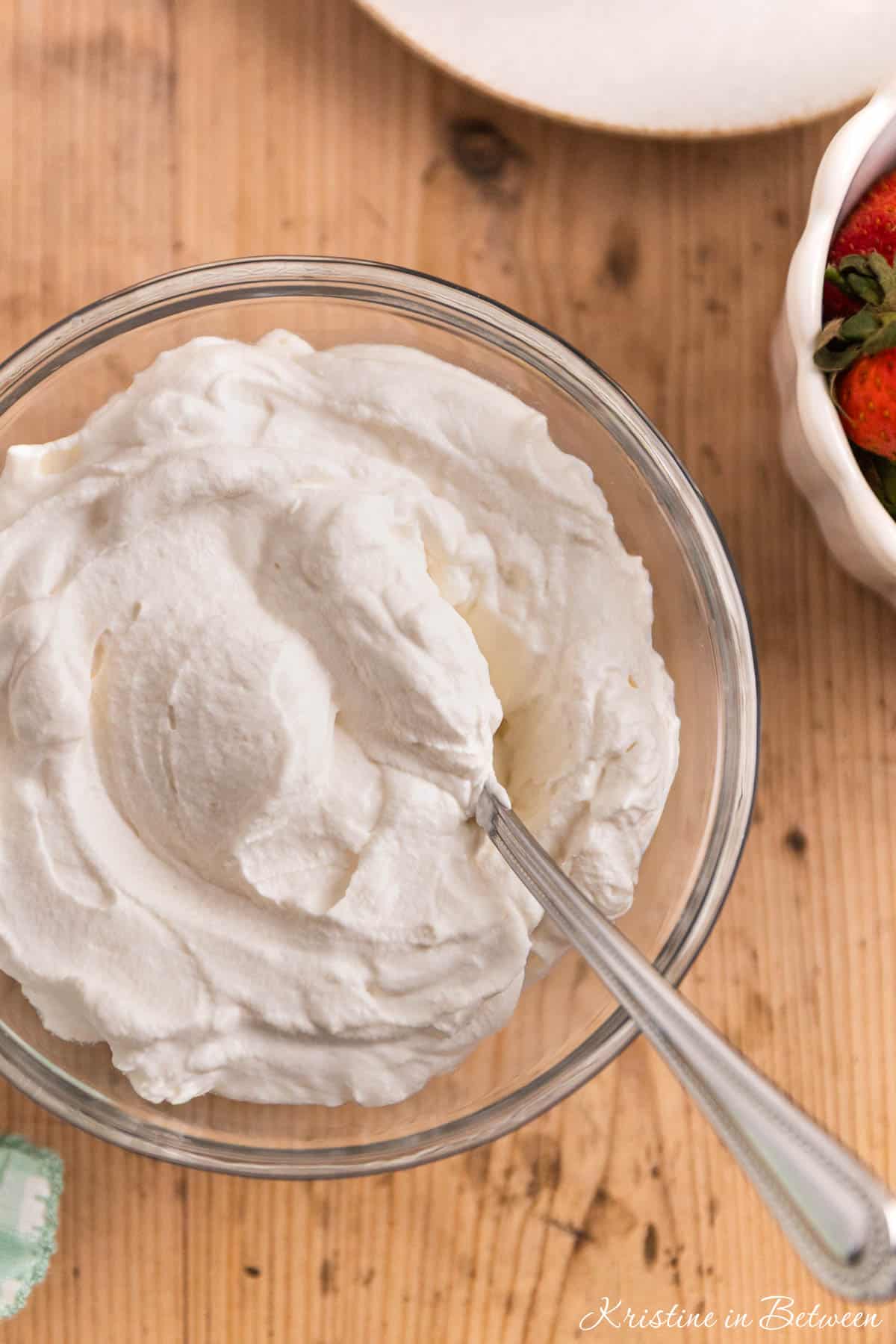 A bowl of homemade whipped cream in a glass bowl with a spoon sitting next to small bowls of berries.