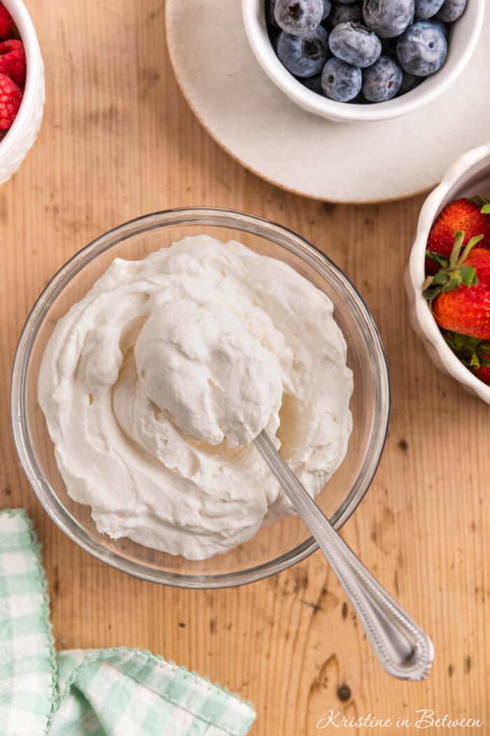 A bowl of homemade whipped cream in a glass bowl with a spoon sitting next to small bowls of berries.