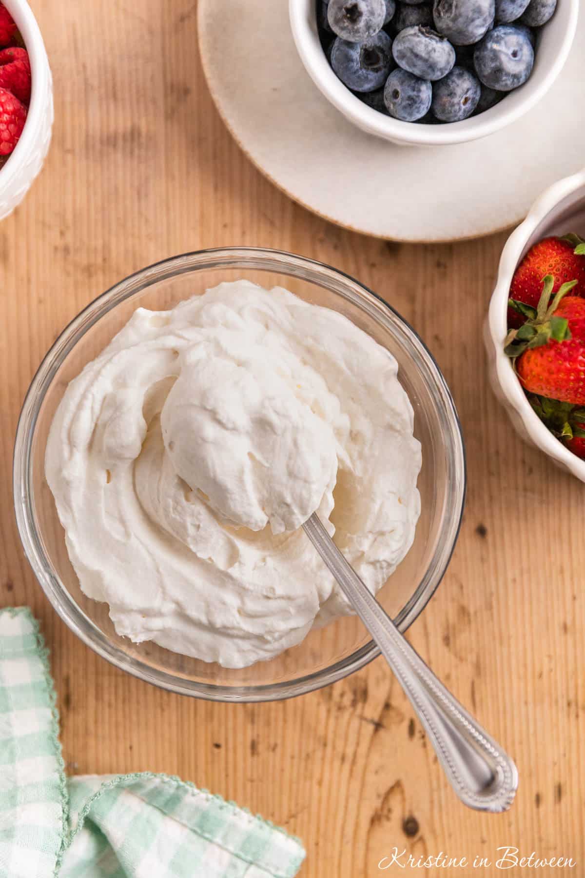 A bowl of homemade whipped cream in a glass bowl with a spoon sitting next to small bowls of berries.