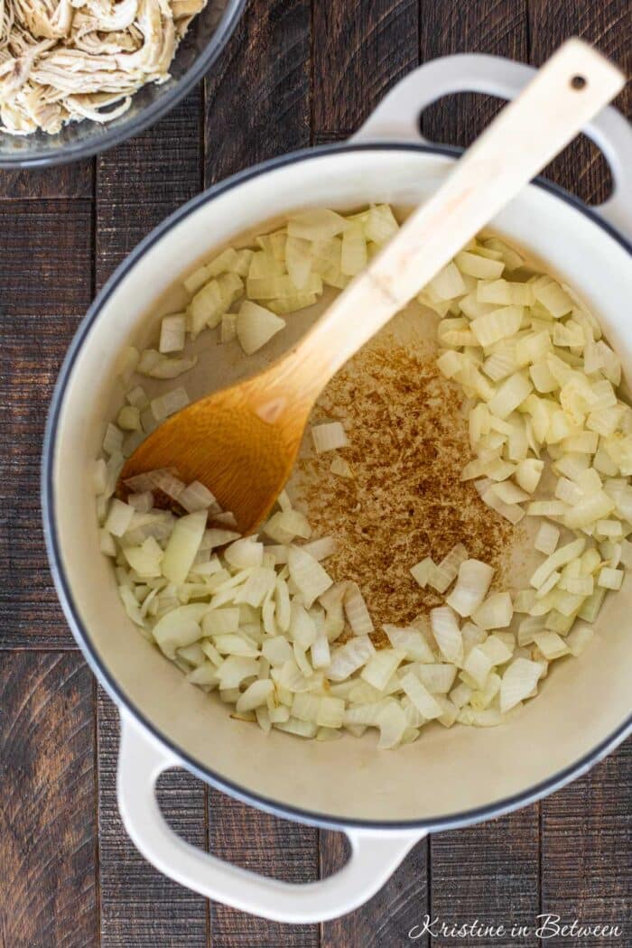 Chopped onion in a Dutch oven with a bowl of shredded chicken in the background.