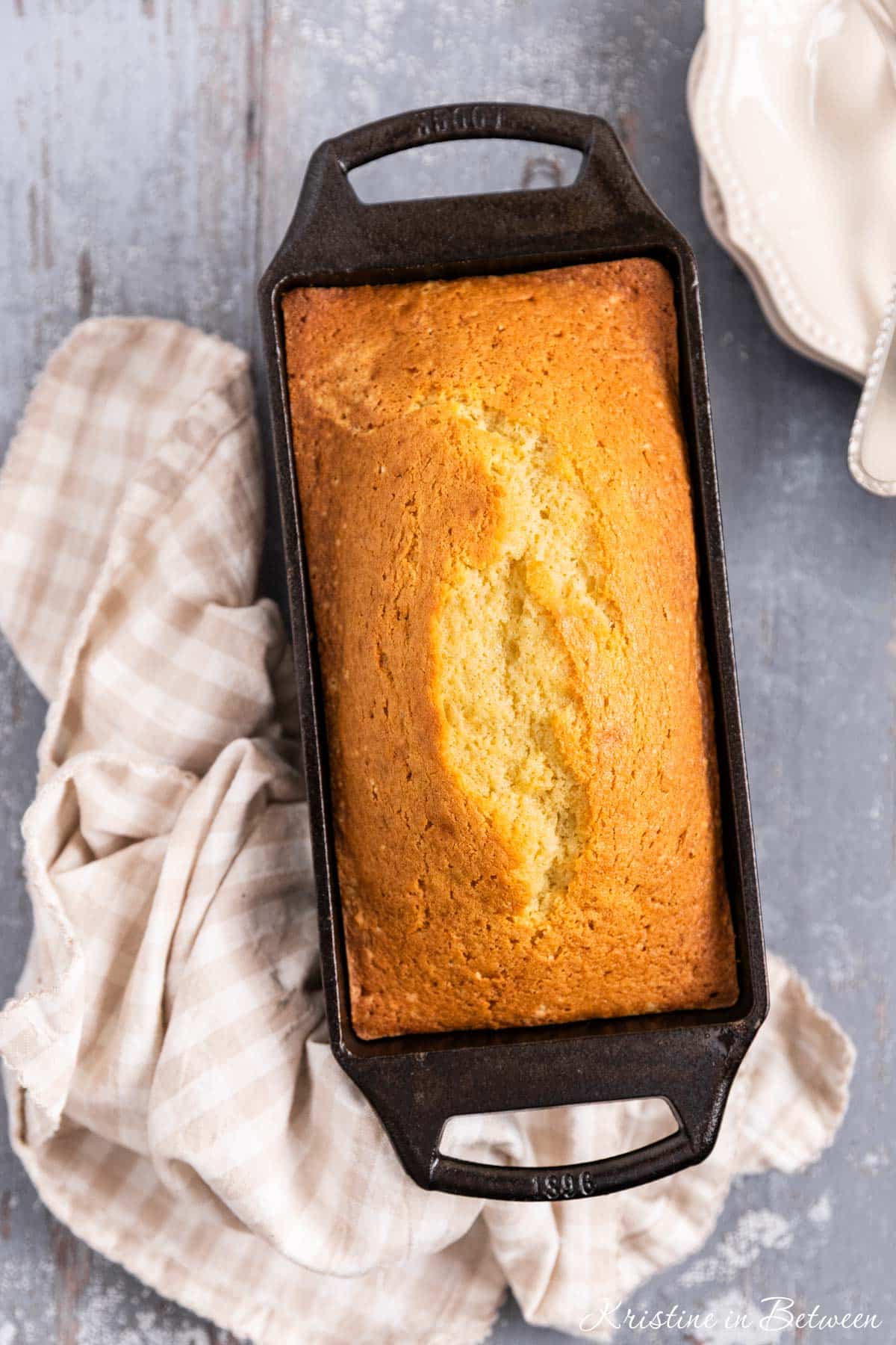 A loaf of pound cake with a pretty crack on top in a cast iron loaf pan with a gingham napkin next to it.