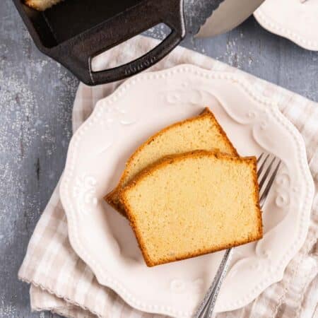 Two slices of old-fashioned pound cake sitting on a white plate with a fork and the rest of the loaf in the background.