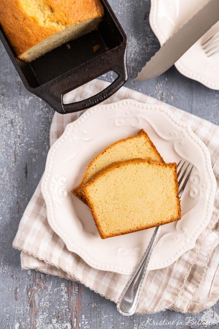 Two slices of old-fashioned pound cake sitting on a white plate with a fork and the rest of the loaf in the background.