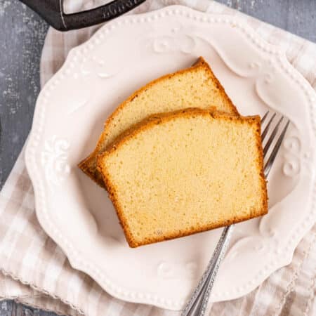 Two slices of old-fashioned pound cake sitting on a white plate with a fork and the rest of the loaf in the background.