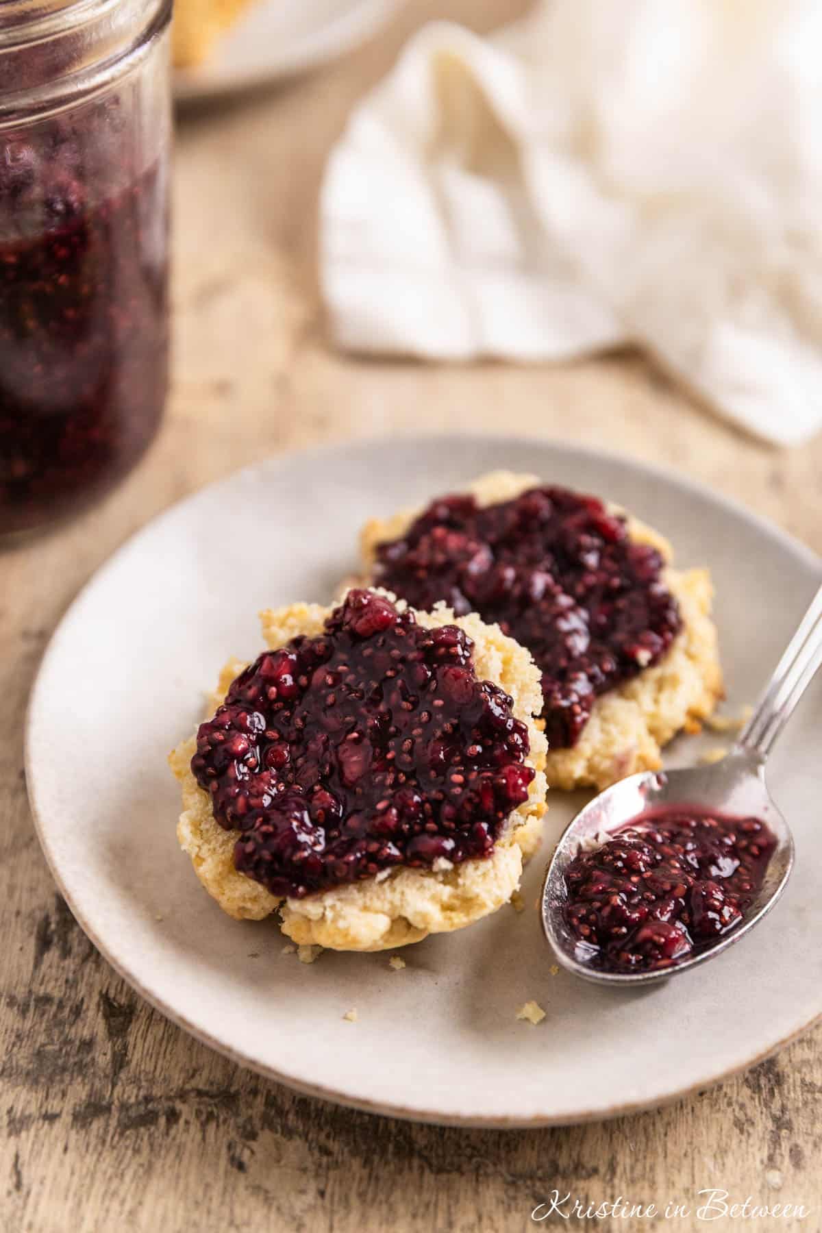 A biscuit cut in half with raspberry chia seed jam spread over the top and a spoon with jam sitting next to it.