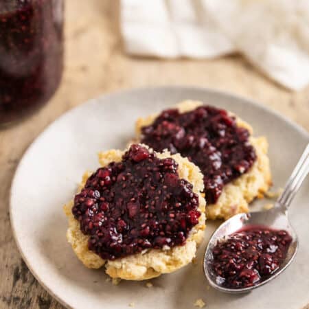A biscuit cut in half with raspberry chia seed jam spread over the top and a spoon with jam sitting next to it.