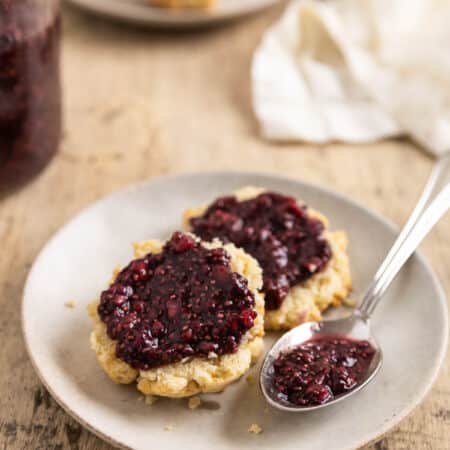A biscuit cut in half with raspberry chia seed jam spread over the top and a spoon with jam sitting next to it.