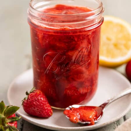 A mason jar of strawberry compote with a spoon laying next to it and a lemon in the background.