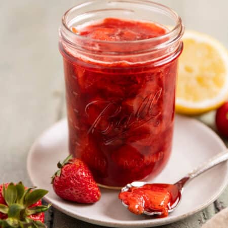 A mason jar of strawberry compote with a spoon laying next to it and a lemon in the background.