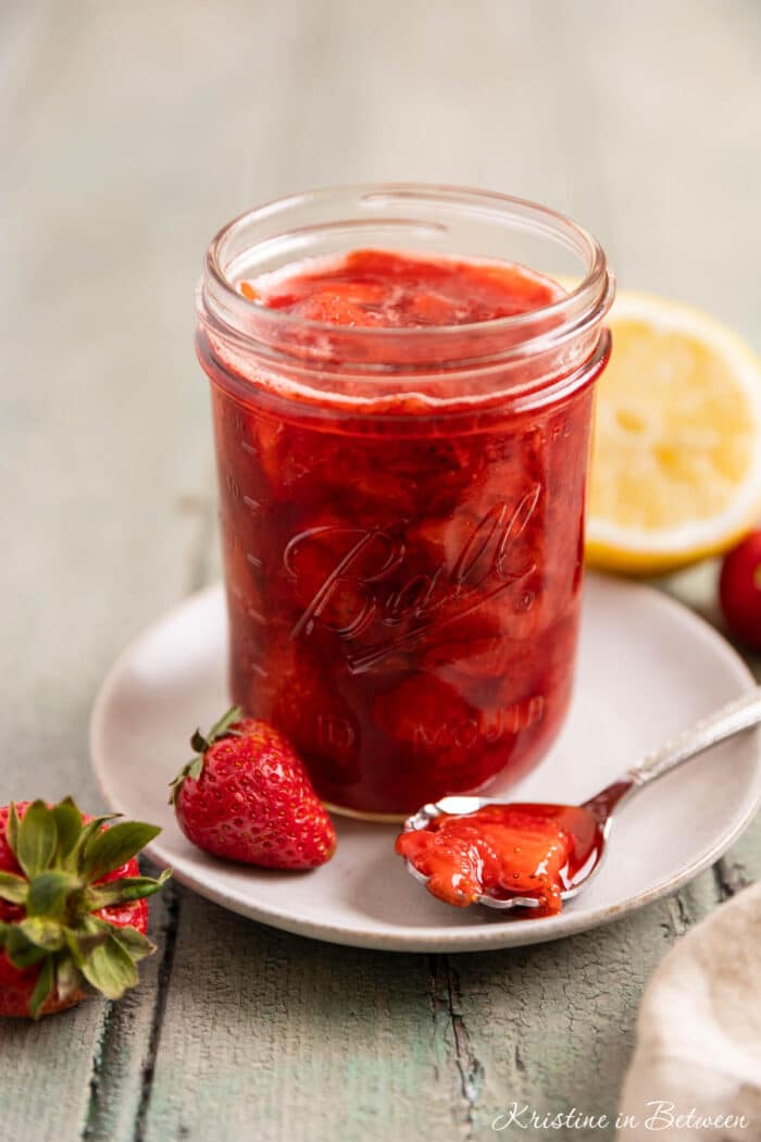 A mason jar of strawberry compote with a spoon laying next to it and a lemon in the background.