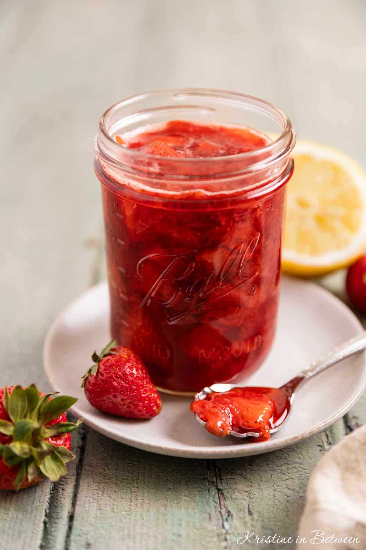 A mason jar of strawberry compote with a spoon laying next to it and a lemon in the background.