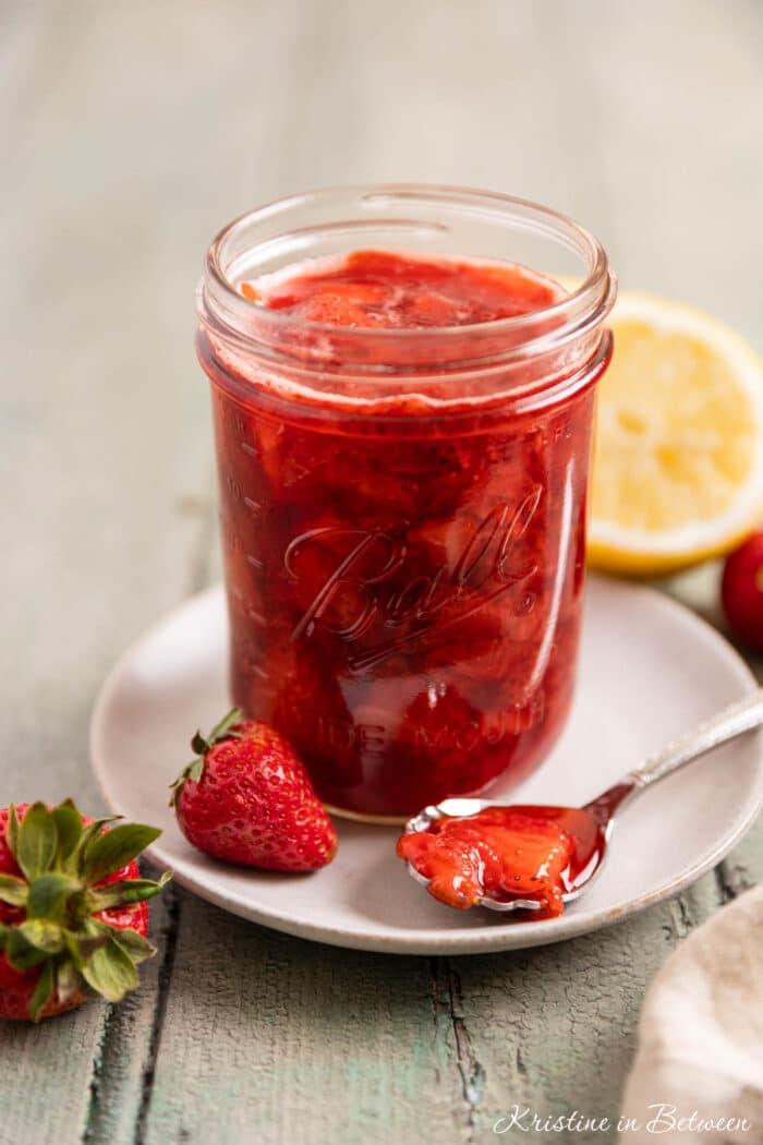 A mason jar of strawberry compote with a spoon laying next to it and a lemon in the background.