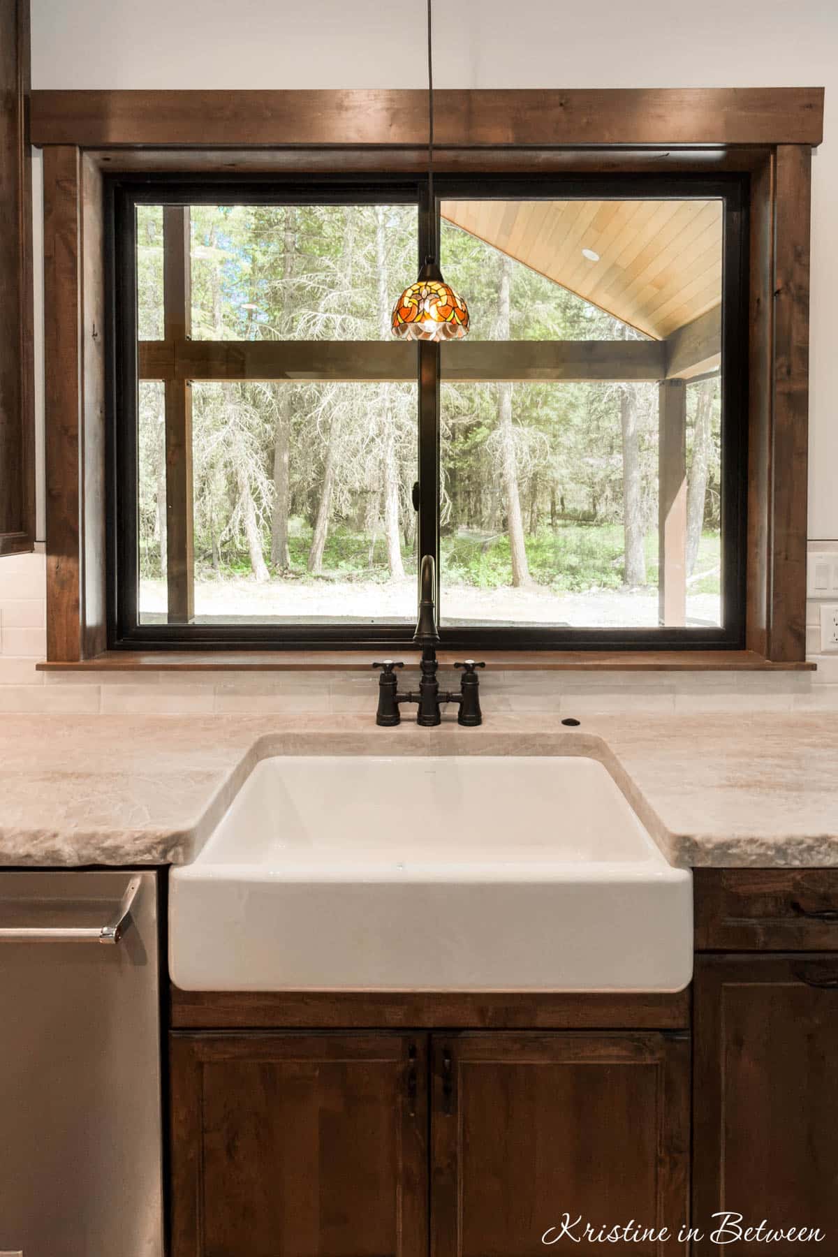 A fancy pendant hanging over a farmhouse sink in front of a window with a view of pine trees.