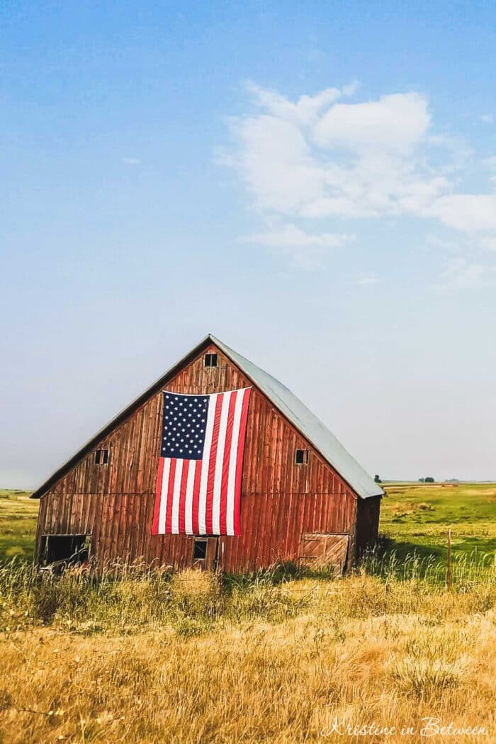 An old red barn in a grassy field with an American flag hanging on the front of it.