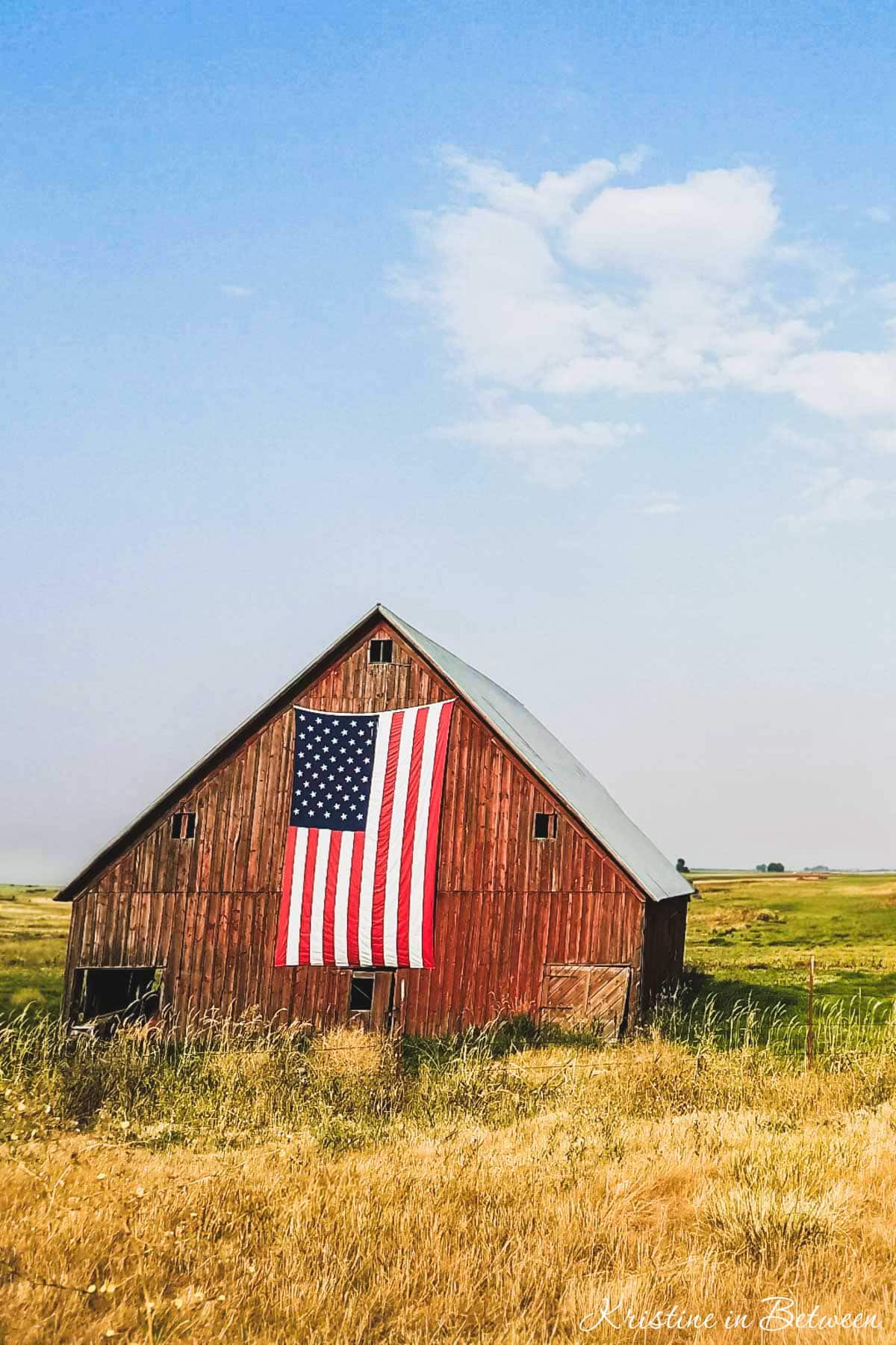 An old red barn in a grassy field with an American flag hanging on the front of it.