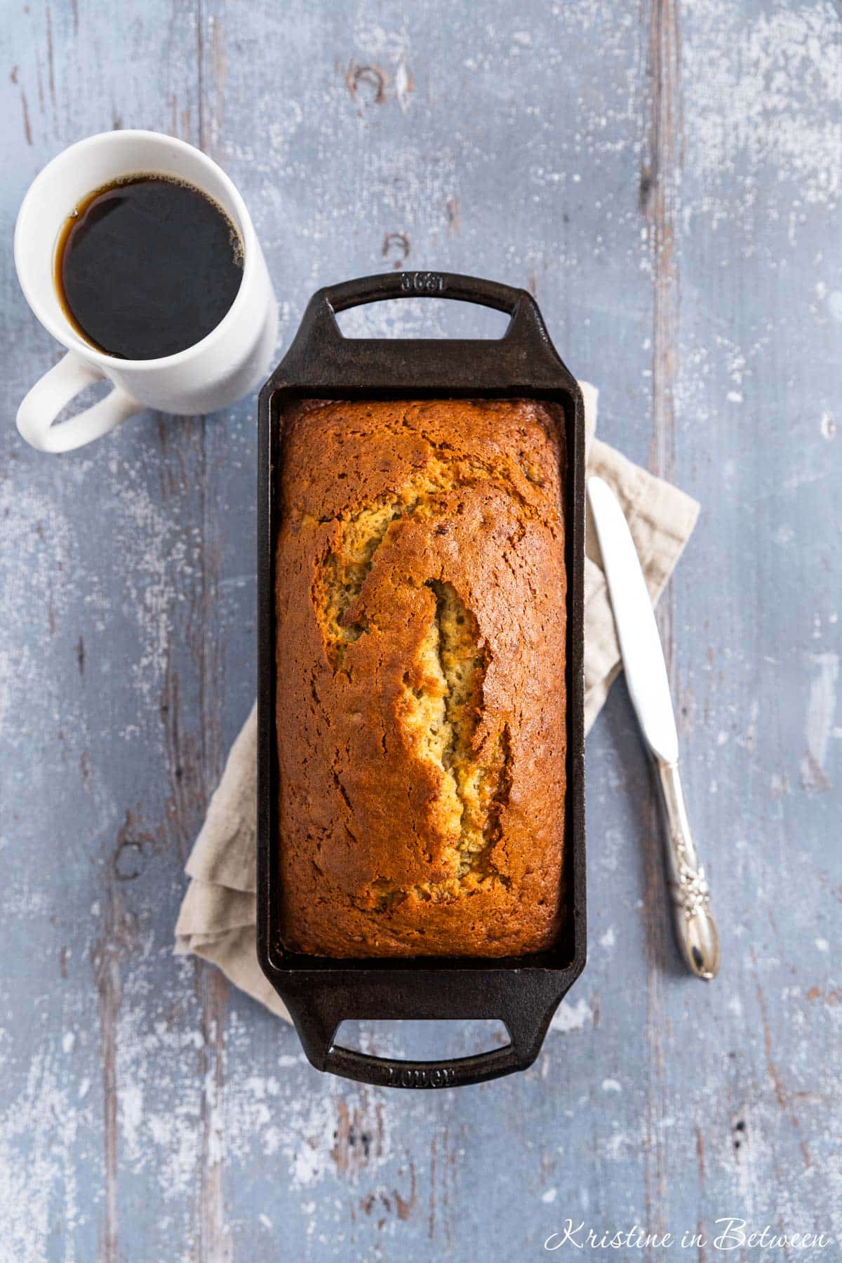 A baked loaf of old-fashioned banana bread in a cast iron loaf pan with a cup of coffee next to it.