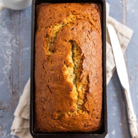 A baked loaf of old-fashioned banana bread in a cast iron loaf pan with a cup of coffee next to it.