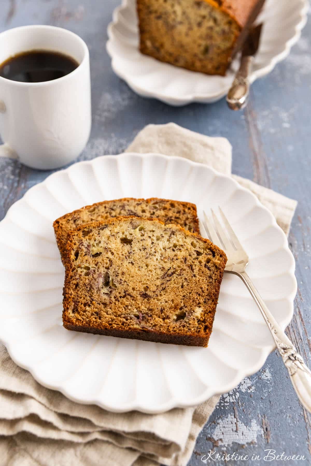 Two slice of banana bread on a white plate with a fork and a cup of coffee in the background.